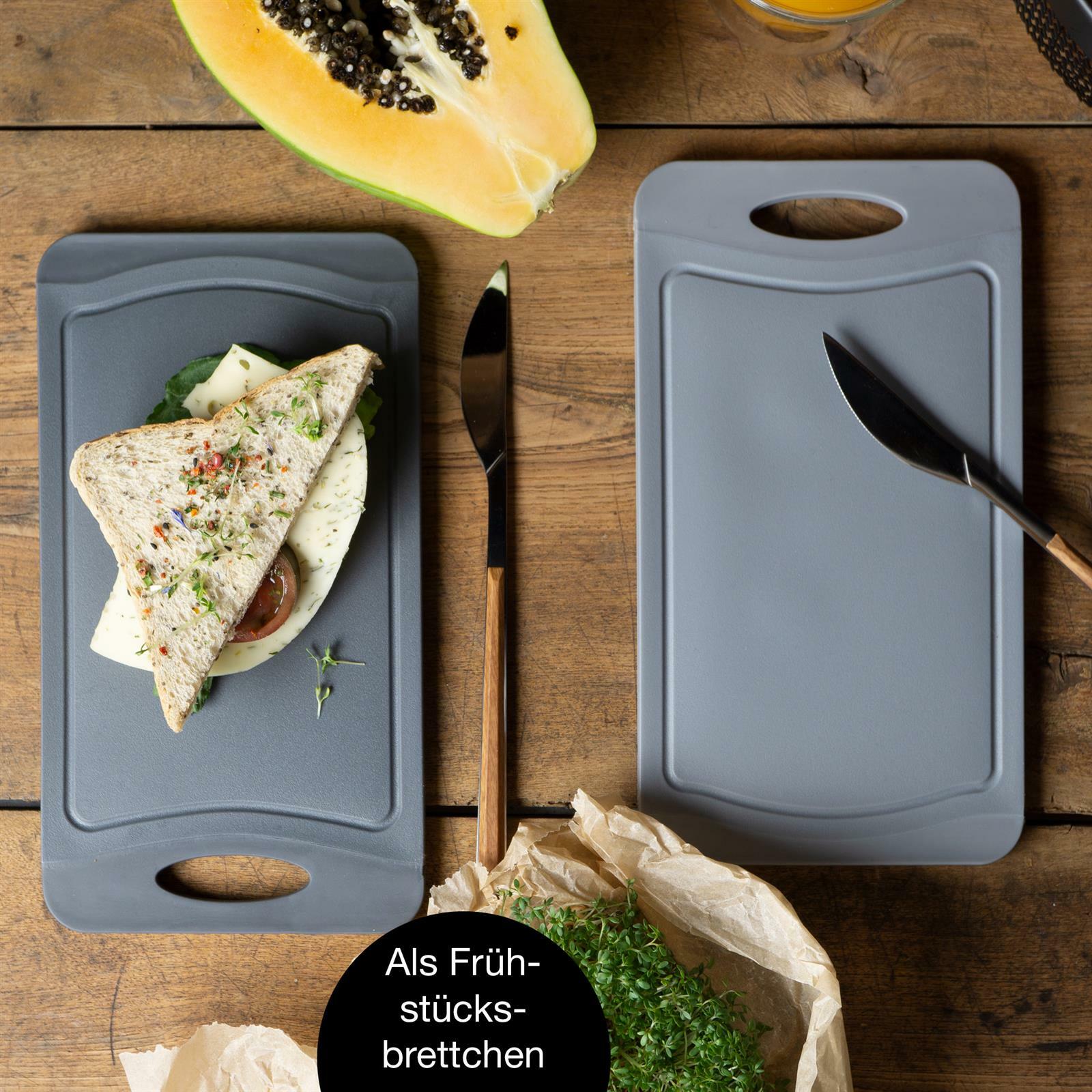 Zwei graue Schneidebrettchen auf einem Holztisch, eines mit einem belegten Sandwich und das andere mit einem Messer darauf. Perspektive von oben.