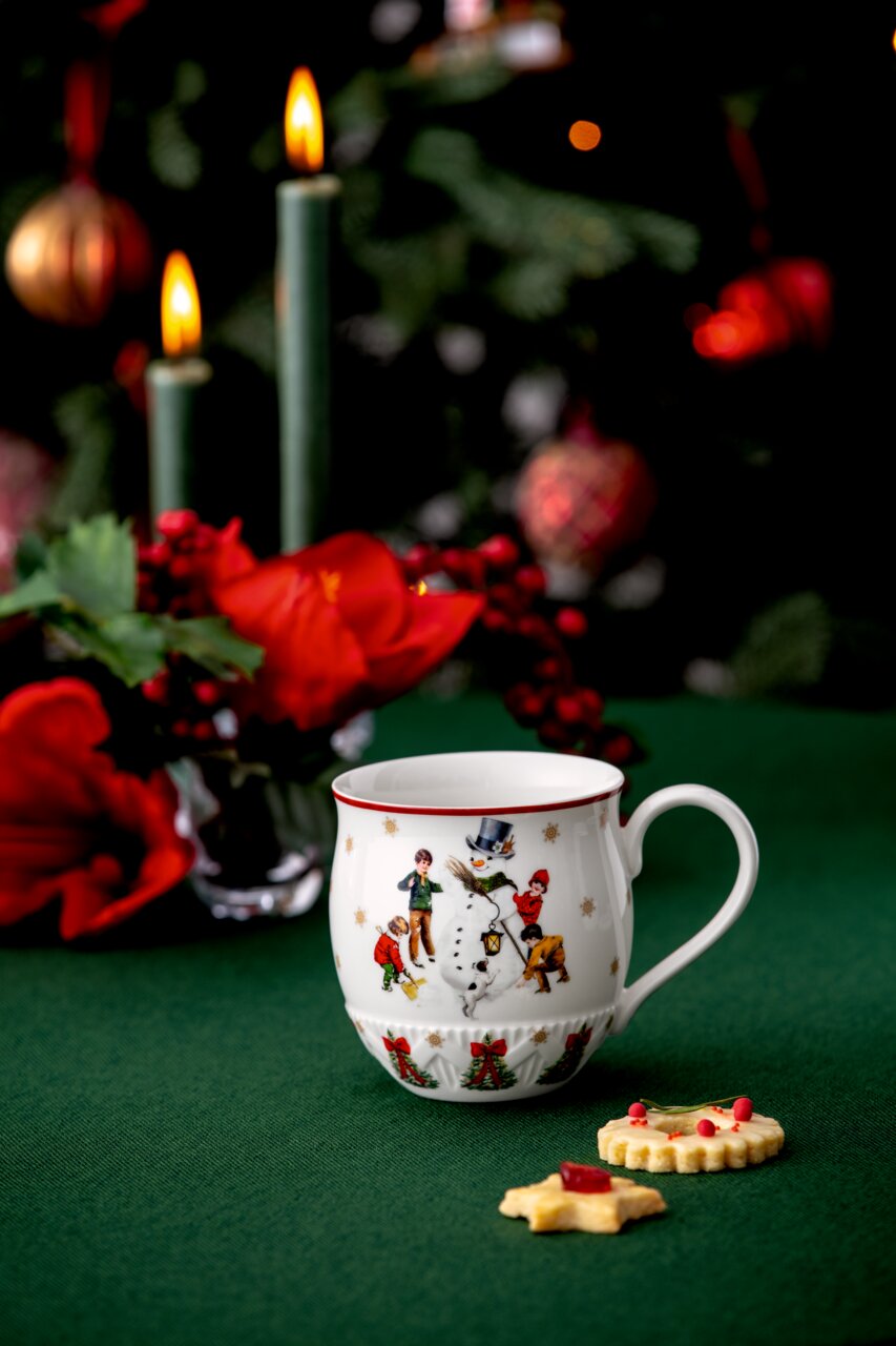 Weihnachtlicher Henkelbecher mit Schneemann-Motiv, umgeben von festlicher Dekoration, aus seitlicher Perspektive fotografiert.