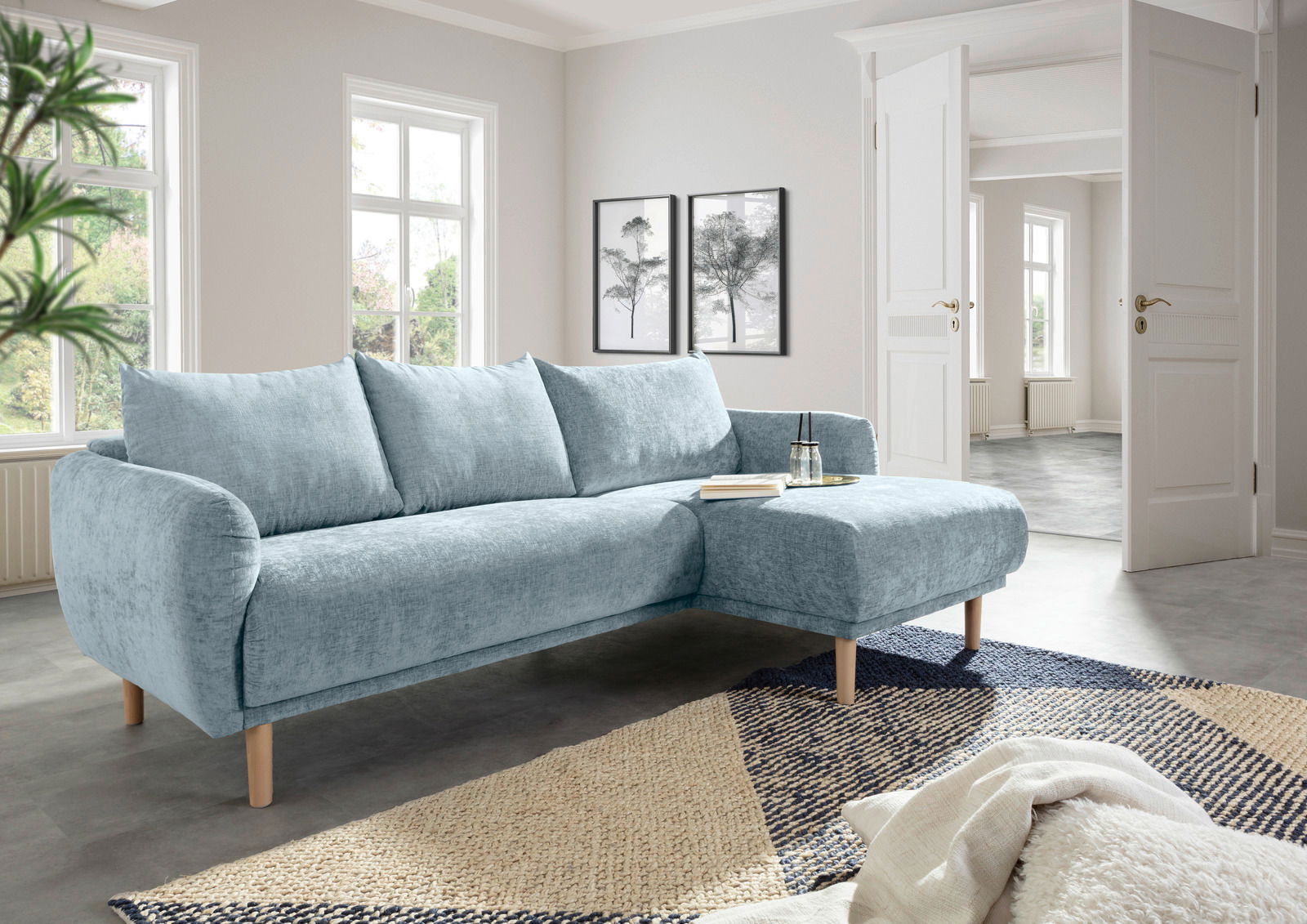 Helles, modernes Ecksofa in einem Wohnzimmer mit großen Fenstern, aus hellblauem Stoff und Holzbeinen, von vorne rechts betrachtet.