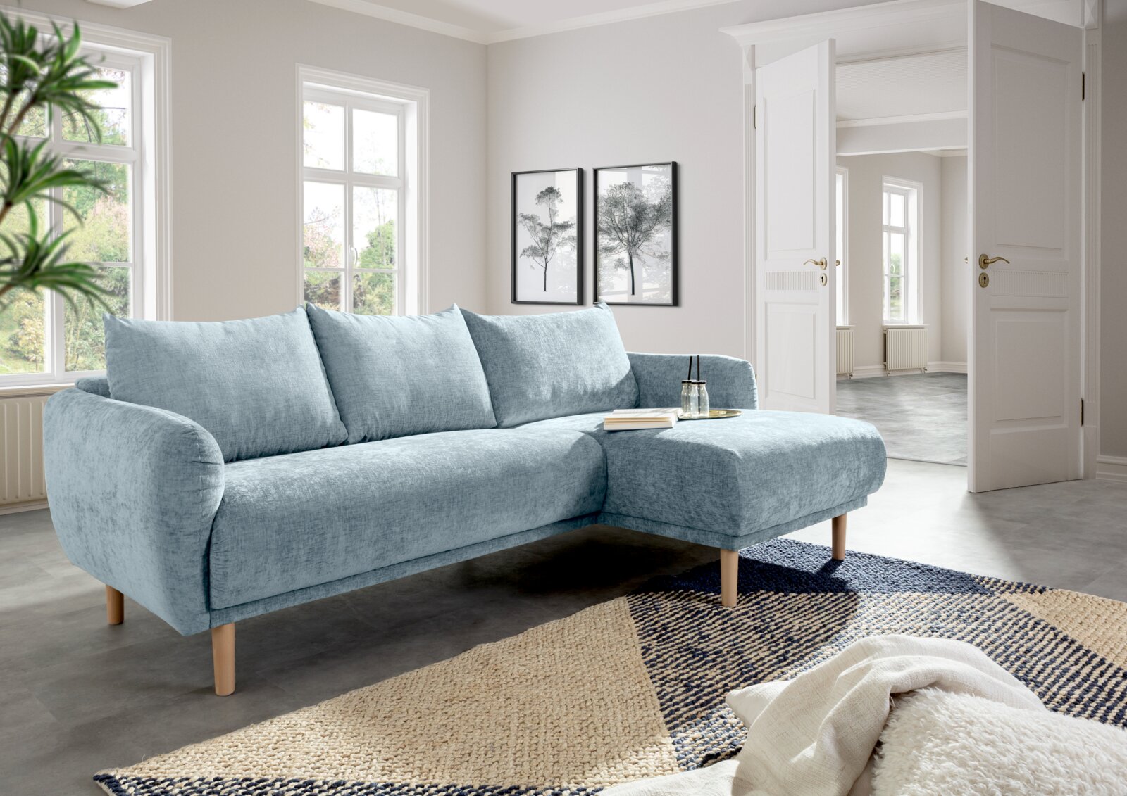 Helles, modernes Ecksofa in einem Wohnzimmer mit großen Fenstern, aus hellblauem Stoff und Holzbeinen, von vorne rechts betrachtet.