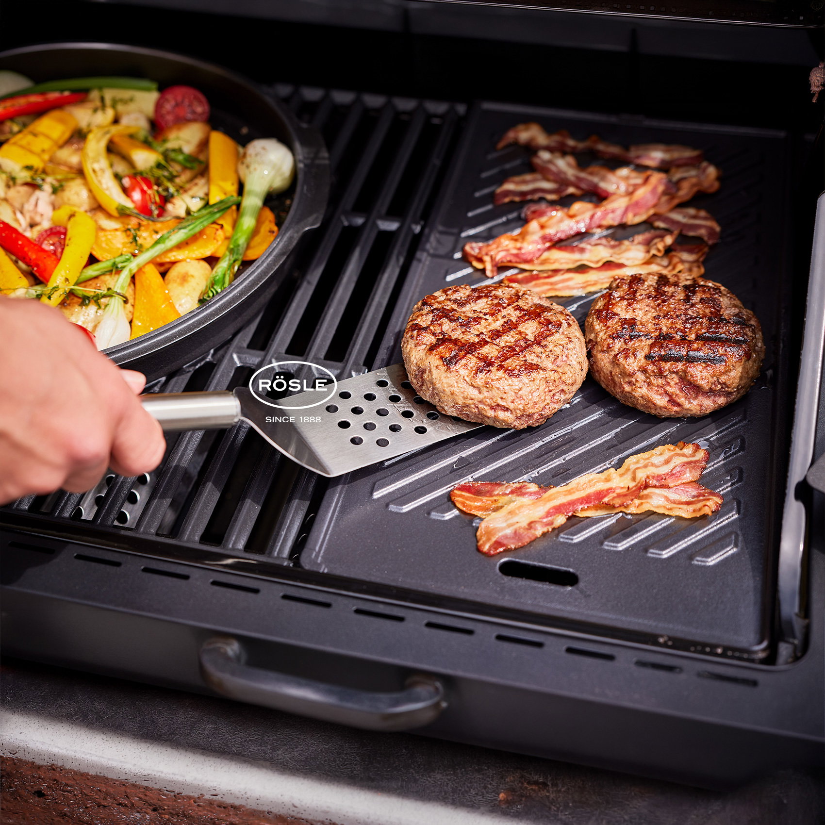 Burger-Patties und Speck auf Grill, daneben Pfanne mit Gemüse, Hand hält Grillspatel.