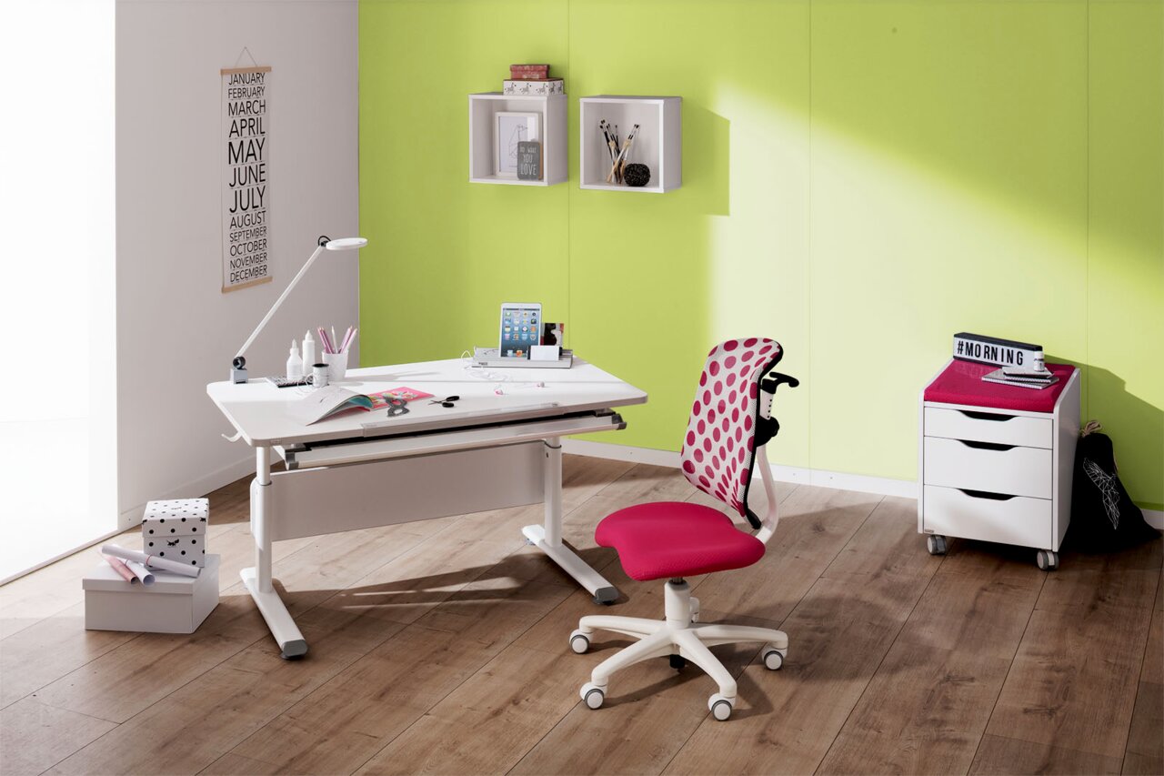 Moderner Schreibtisch mit weißer Oberfläche und verstellbarer Höhe, begleitet von einem pink gepunkteten Jugendstuhl, in einem hellen Raum mit grüner Wand, aus seitlicher Perspektive.