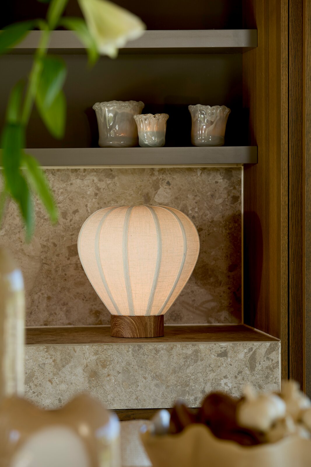 Leuchtende Tischlampe mit ballonförmigem, cremefarbenem Stoffschirm und Holzsockel, frontal in einer eleganten Naturstein- und Holznische inszeniert