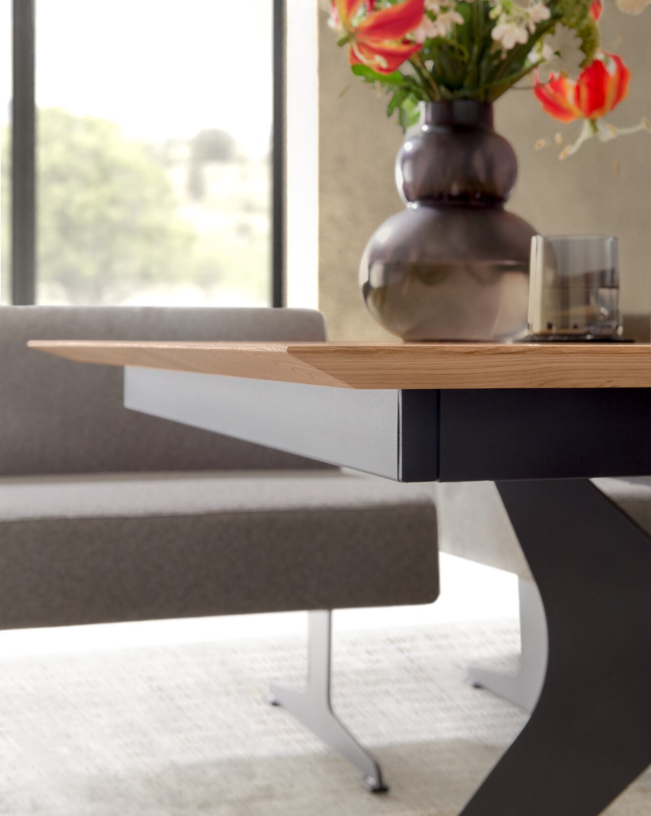 Detailansicht eines modernen Esstisches aus hellem Holz mit schwarzem Metallgestell in X-Form, seitliche Perspektive auf Tischkante und Untergestell, im Hintergrund eine gepolsterte Bank sowie Vase mit Blumen und Glas auf der Tischplatte.