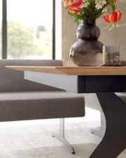 Detailansicht eines modernen Esstisches aus hellem Holz mit schwarzem Metallgestell in X-Form, seitliche Perspektive auf Tischkante und Untergestell, im Hintergrund eine gepolsterte Bank sowie Vase mit Blumen und Glas auf der Tischplatte.