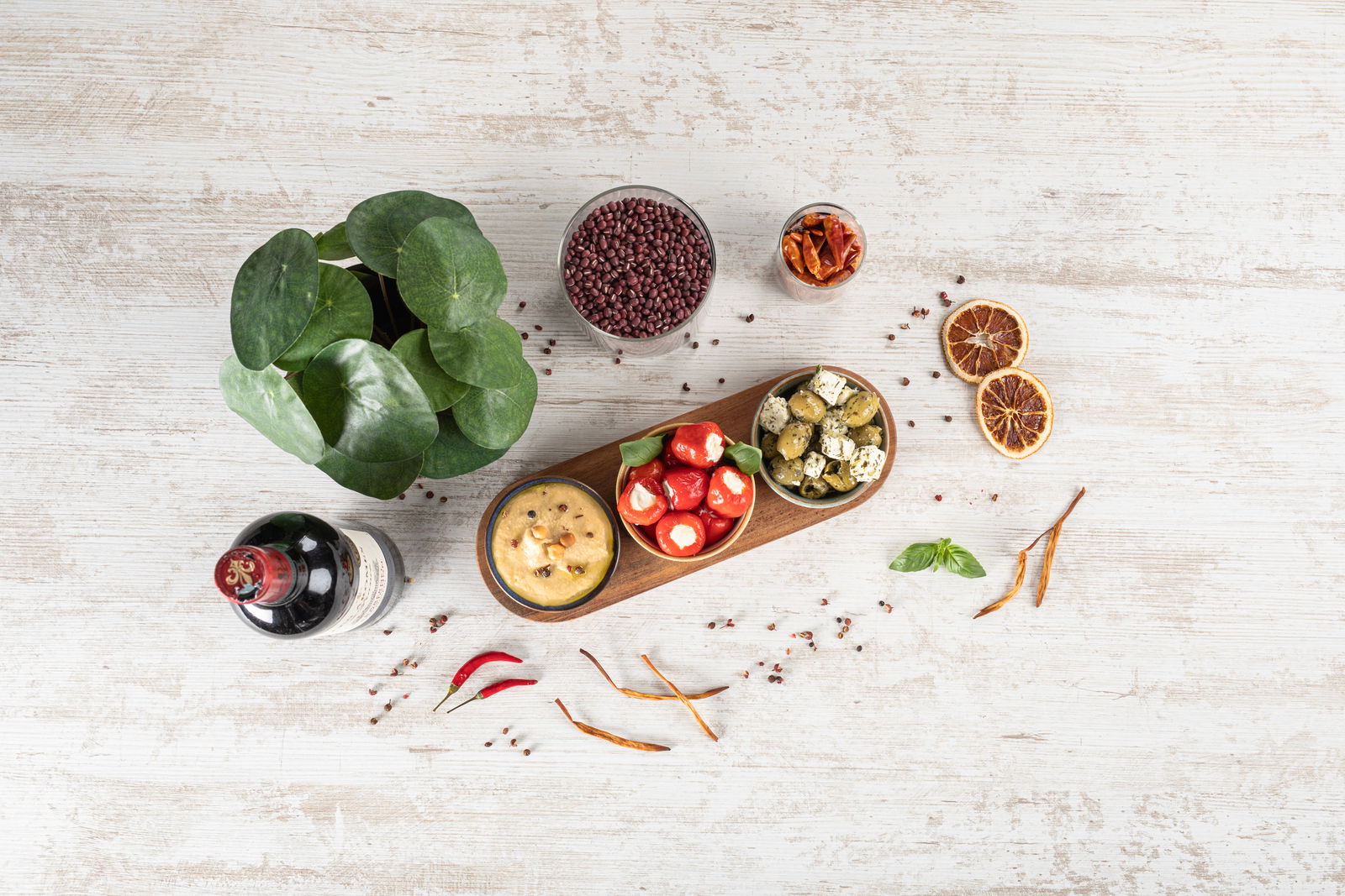 Vogelperspektive auf ein Akazienbrett mit drei runden Schalen, gefüllt mit verschiedenen Antipasti, umgeben von dekorativen Elementen wie einer Pflanze, einer Flasche Wein und getrockneten Früchten.