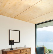 Moderne Tischleuchte mit schwarzem, gemustertem Lampenschirm auf einer Holzkommode, seitliche Perspektive mit Blick auf ein großes Fenster und Berglandschaft.