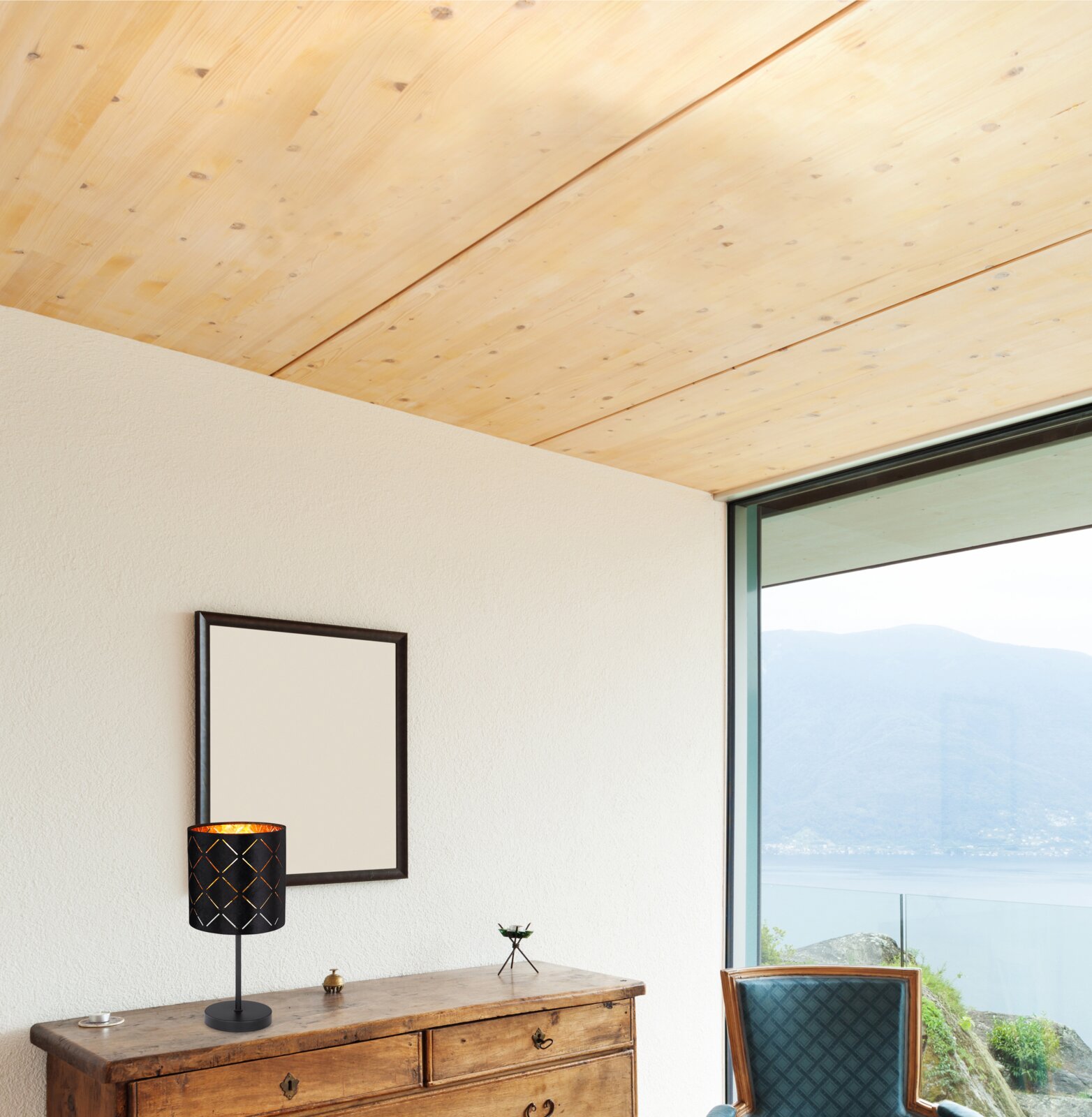 Moderne Tischleuchte mit schwarzem, gemustertem Lampenschirm auf einer Holzkommode, seitliche Perspektive mit Blick auf ein großes Fenster und Berglandschaft.