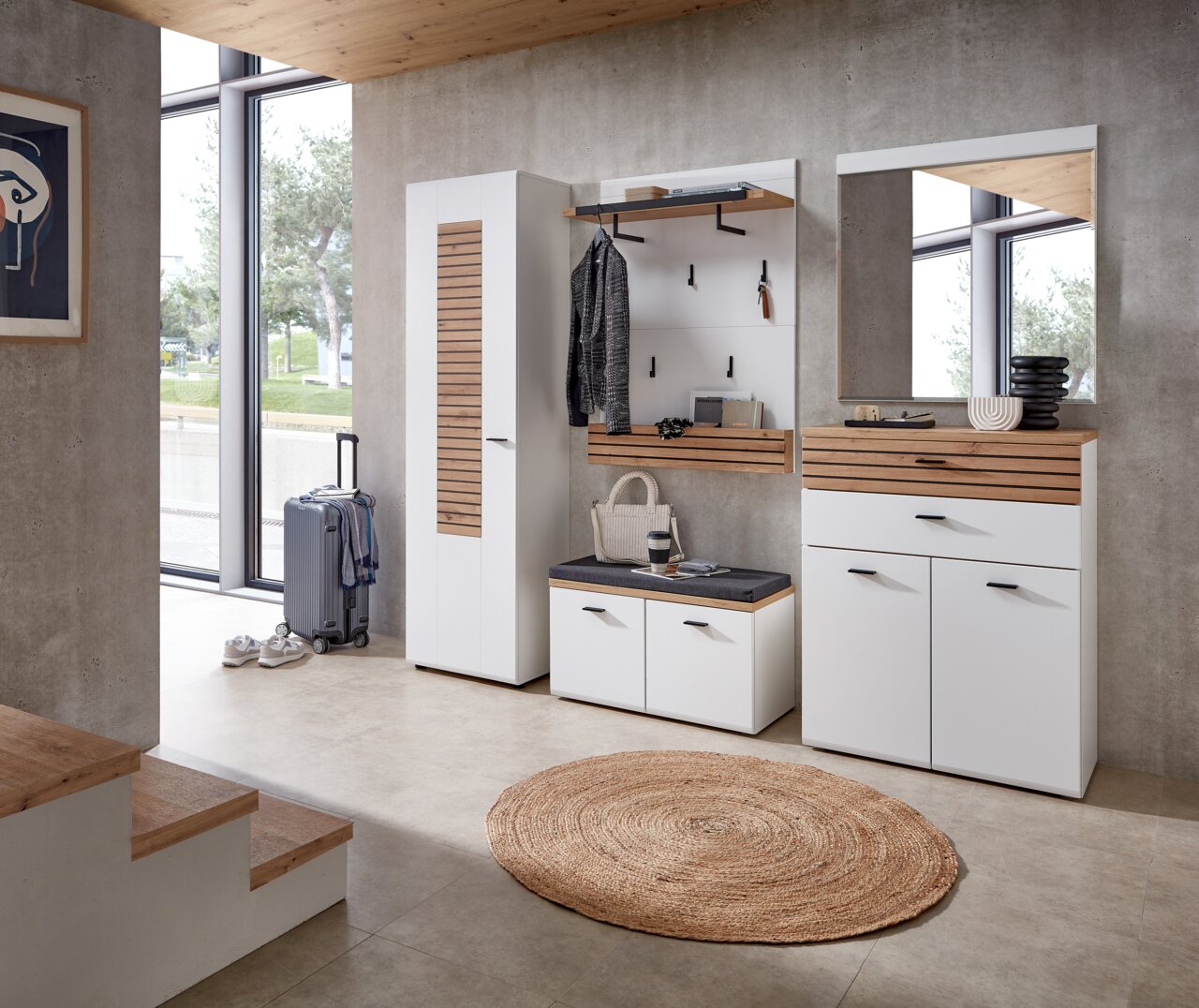 Moderne Garderobenkombination in einem hellen Raum mit großen Fenstern, bestehend aus einem hohen Schrank, einer Sitzbank mit Stauraum, einer Wandgarderobe mit Haken und einem Spiegel, aus der Perspektive des Raumes betrachtet.