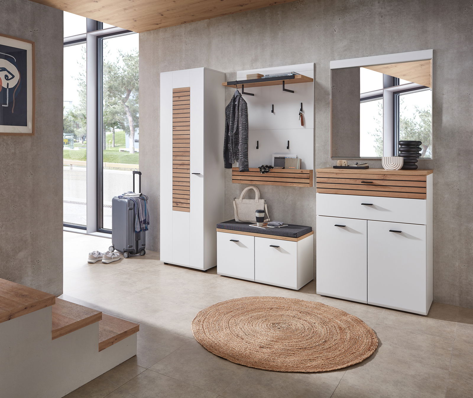 Moderne Garderobenkombination in einem hellen Raum mit großen Fenstern, bestehend aus einem hohen Schrank, einer Sitzbank mit Stauraum, einer Wandgarderobe mit Haken und einem Spiegel, aus der Perspektive des Raumes betrachtet.