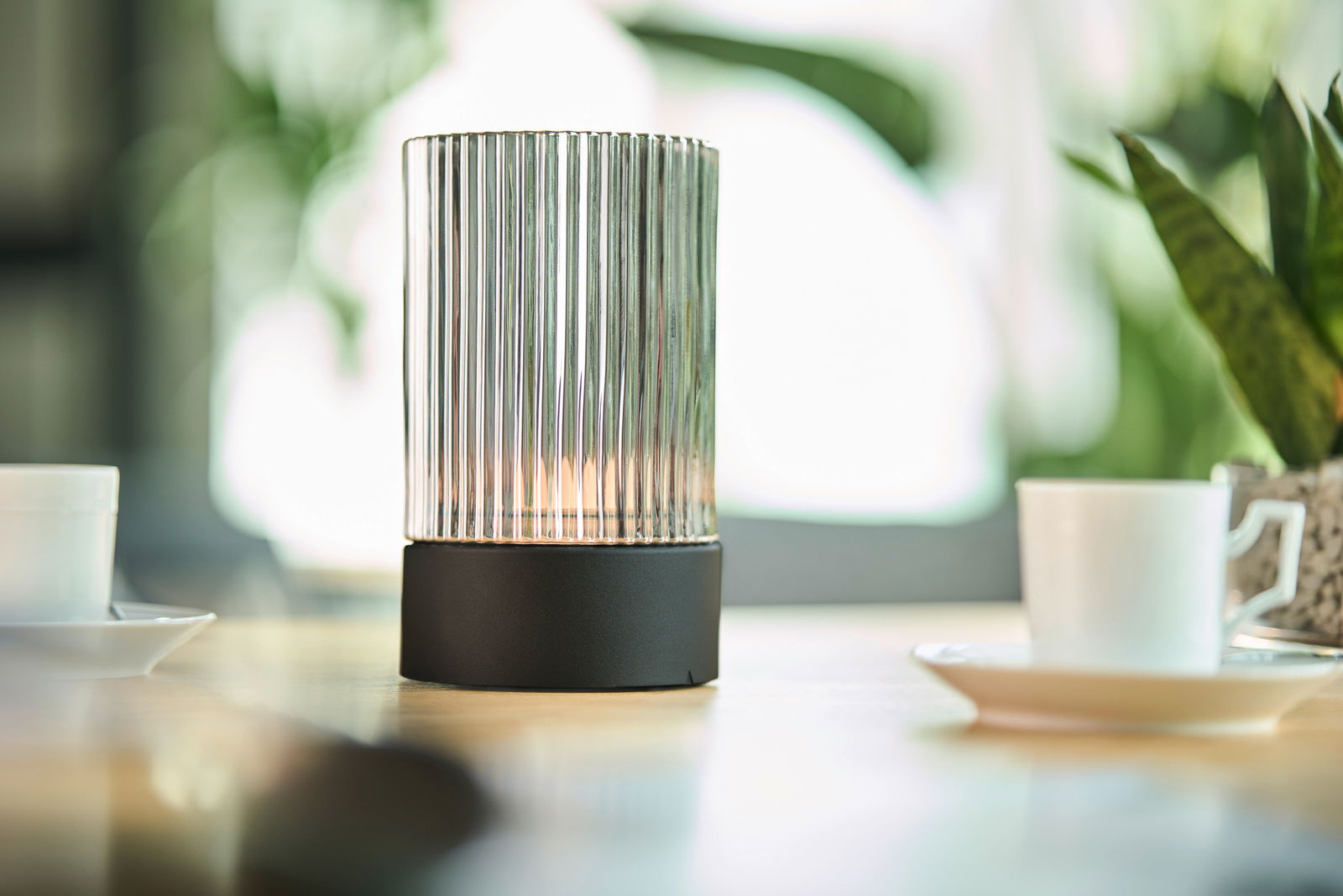 Akku-Windlicht mit geriffeltem Glaszylinder und schwarzem Sockel, auf einem Tisch mit Tassen und Pflanzen im Hintergrund, seitliche Perspektive