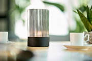 Akku-Windlicht mit geriffeltem Glaszylinder und schwarzem Sockel, auf einem Tisch mit Tassen und Pflanzen im Hintergrund, seitliche Perspektive