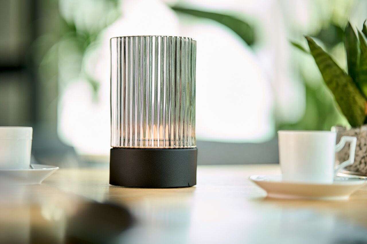 Akku-Windlicht mit geriffeltem Glaszylinder und schwarzem Sockel, auf einem Tisch mit Tassen und Pflanzen im Hintergrund, seitliche Perspektive