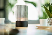 Akku-Windlicht mit geriffeltem Glaszylinder und schwarzem Sockel, auf einem Tisch mit Tassen und Pflanzen im Hintergrund, seitliche Perspektive