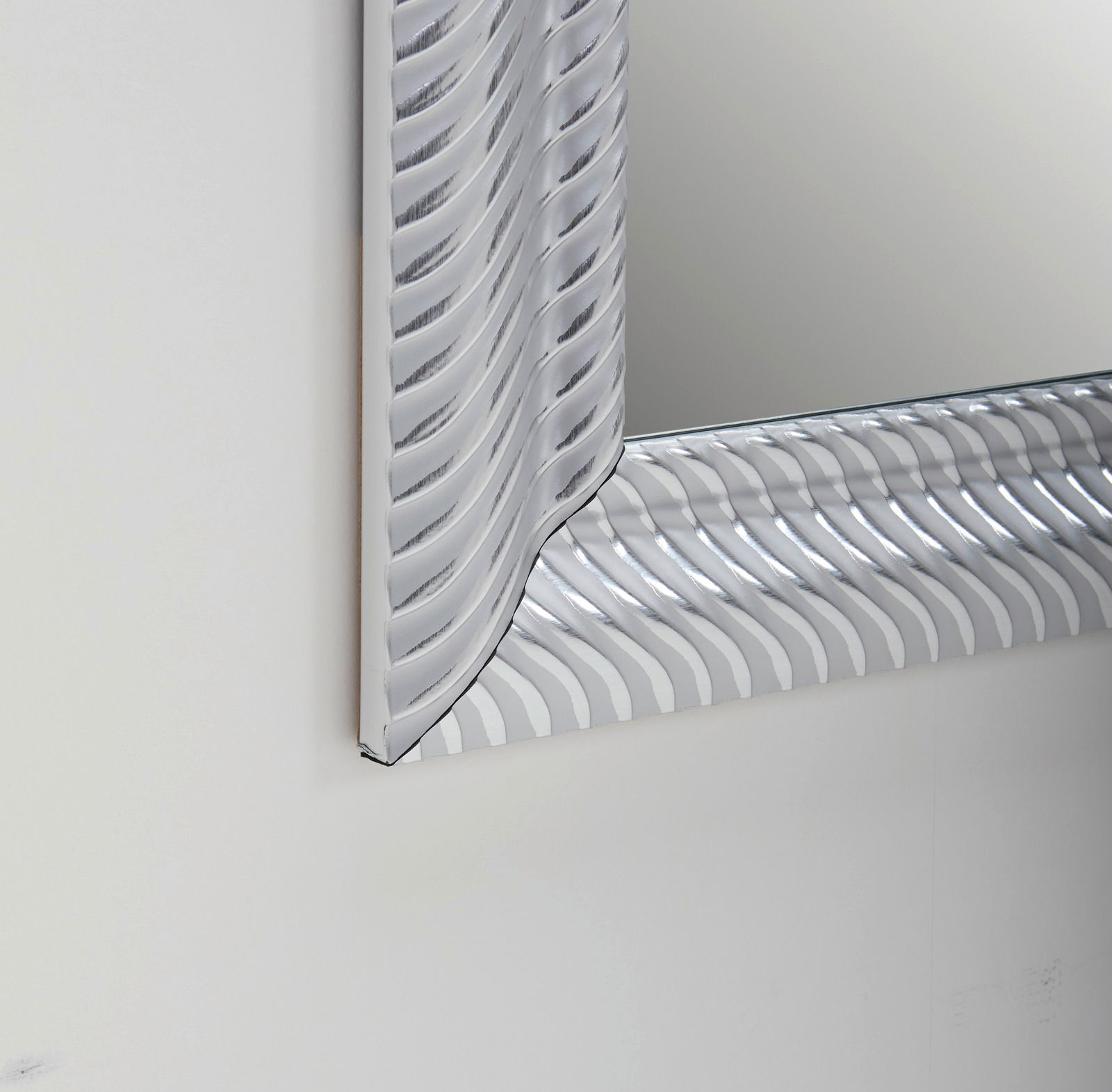 Nahaufnahme eines rechteckigen Spiegels mit dekorativem, wellenförmigem Rahmen in Silberoptik, perspektivisch von der Seite aufgenommen.