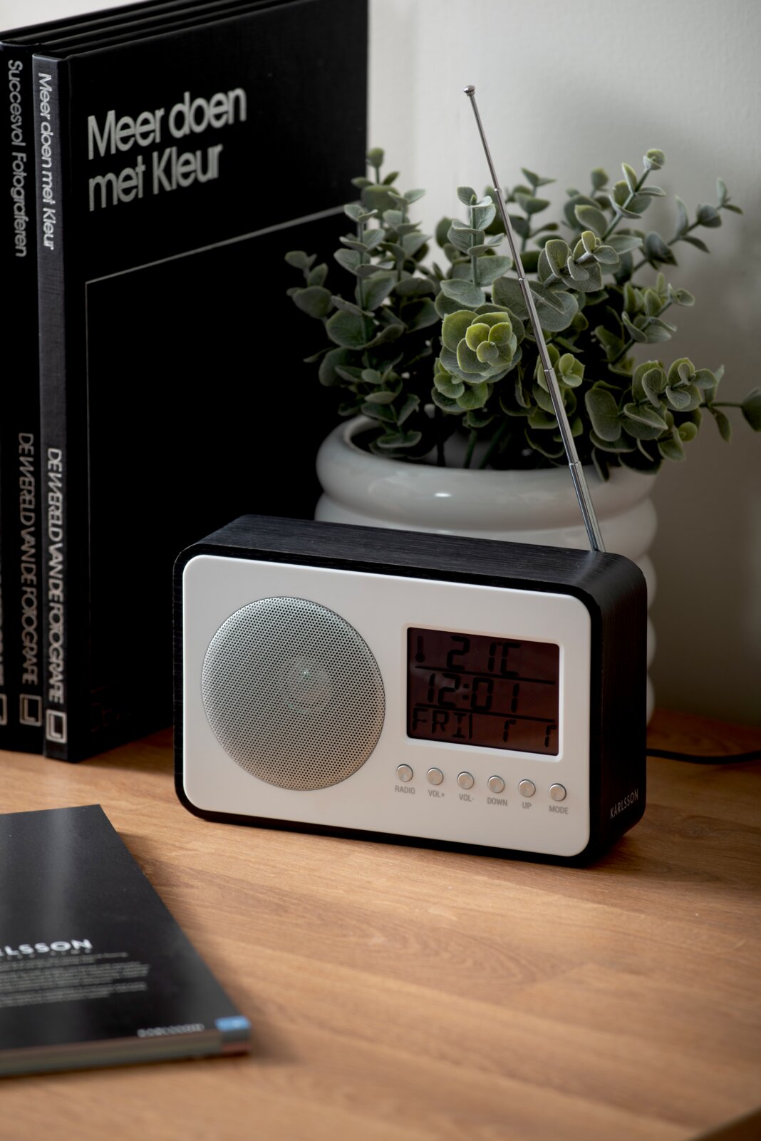 Moderne wekkerradio met digitaal display en antenne, op een tafel naast planten en boeken, gefotografeerd vanuit een zijdelingse hoek.