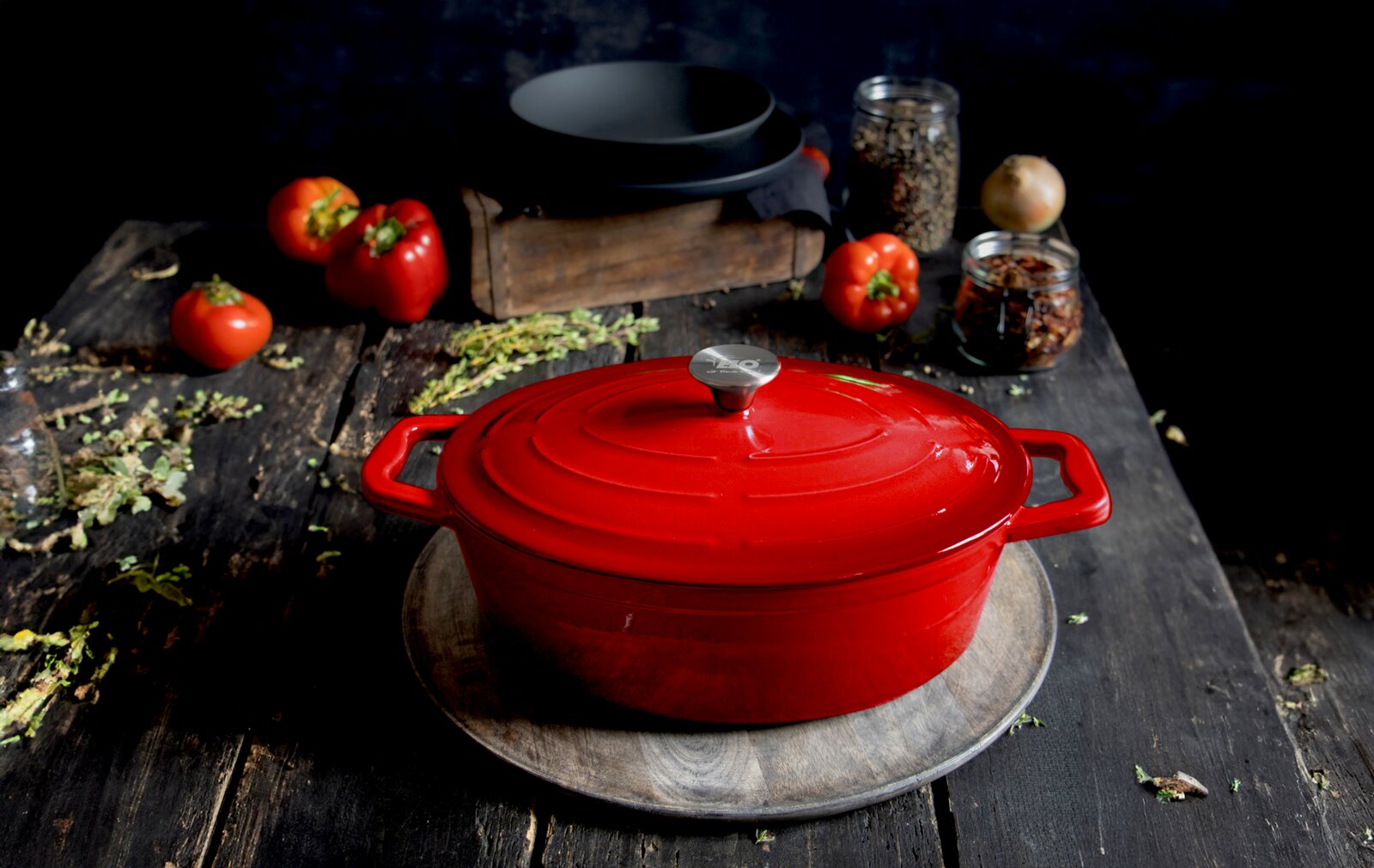 Rode ovale gietijzeren braadpan met deksel, van bovenaf gefotografeerd op een rustieke houten tafel, omringd door verse tomaten en kruiden.
