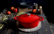 Rode ovale gietijzeren braadpan met deksel, van bovenaf gefotografeerd op een rustieke houten tafel, omringd door verse tomaten en kruiden.