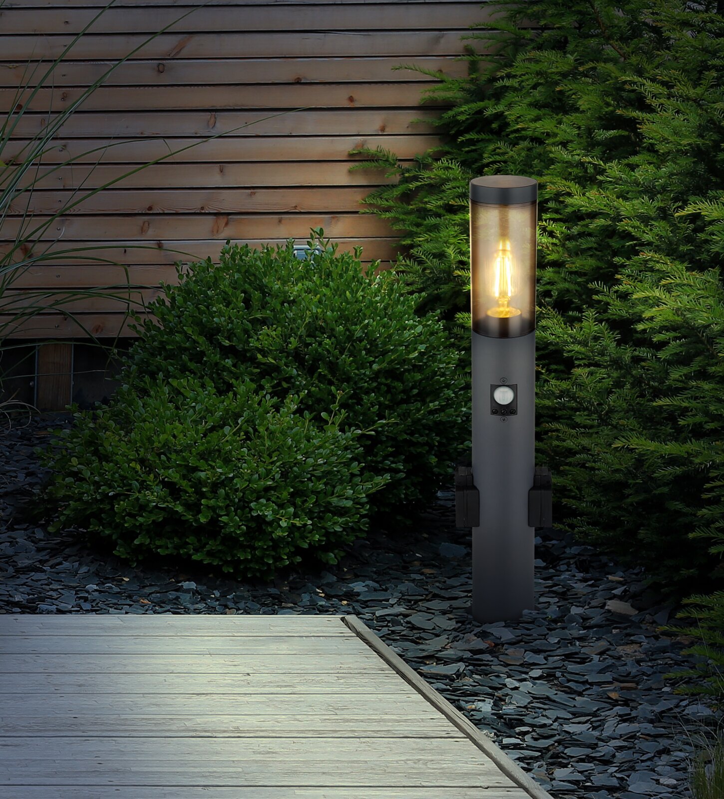 Moderne Außenleuchte in einem Garten, seitliche Perspektive, mit warmem Lichtschein, umgeben von Pflanzen und Holzwand.