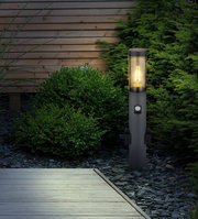 Moderne Außenleuchte in einem Garten, seitliche Perspektive, mit warmem Lichtschein, umgeben von Pflanzen und Holzwand.