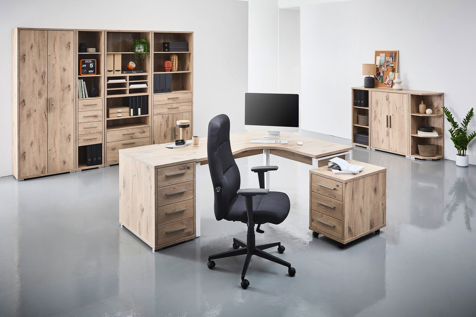 Moderne Büromöbel in Holzoptik, bestehend aus einem Eckschreibtisch mit Rollcontainer und einem ergonomischen Bürostuhl, umgeben von hohen Aktenschränken und Regalen. Perspektive: Frontalansicht.