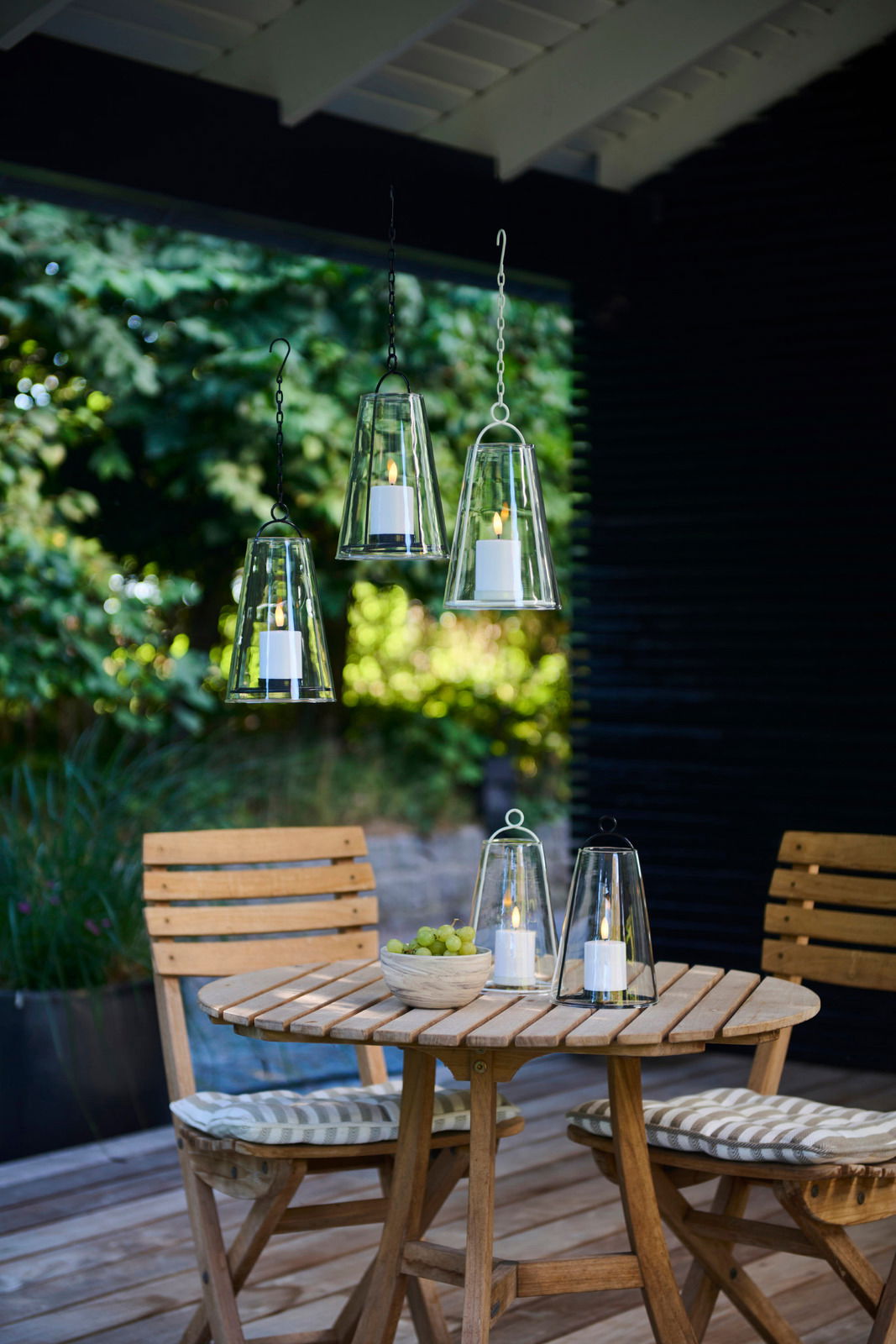 Sirius Windlicht mit LED Kerze ALBERT Hängende und stehende Windlichter mit LED-Kerzen auf einer Terrasse, umgeben von grüner Natur, aus der Perspektive eines Betrachters auf Augenhöhe.