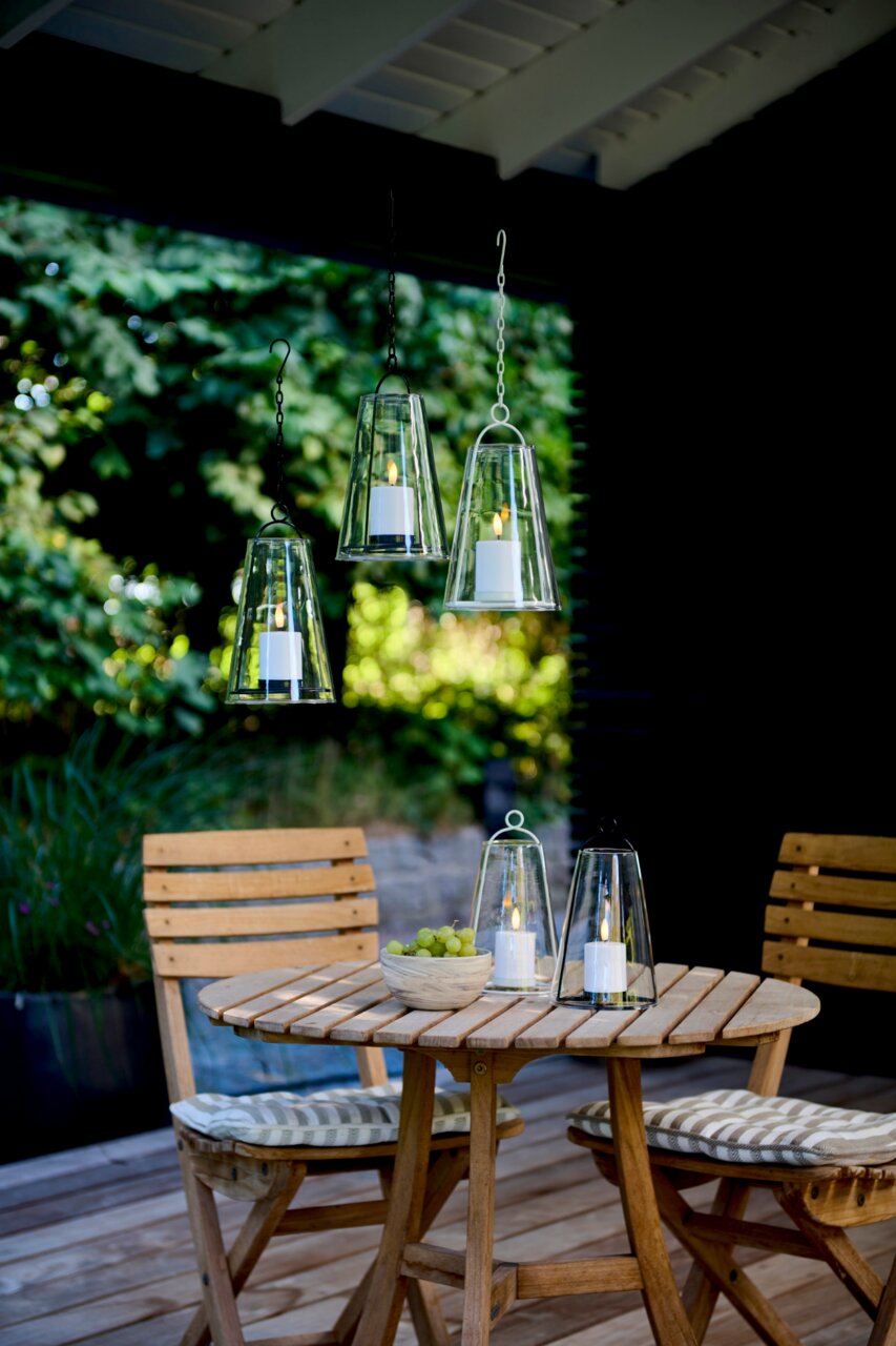 Hangende en staande windlichten met led-kaarsen op een terras, omgeven door groene natuur, gezien vanuit het perspectief van een toeschouwer op ooghoogte.