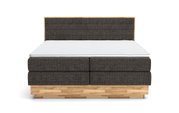Vooraanzicht van een boxspringbed met een donkere stoffen bekleding en een licht houten frame.