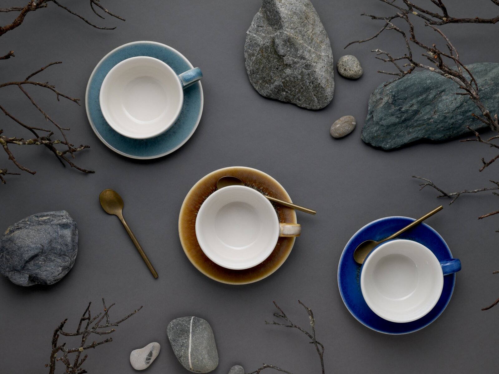 Drei leere Keramik-Kaffeetassen mit passenden Untertassen in Blau- und Brauntönen, dekoriert mit Messinglöffeln, arrangiert zwischen Steinen und Zweigen auf grauem Hintergrund, Draufsicht von oben