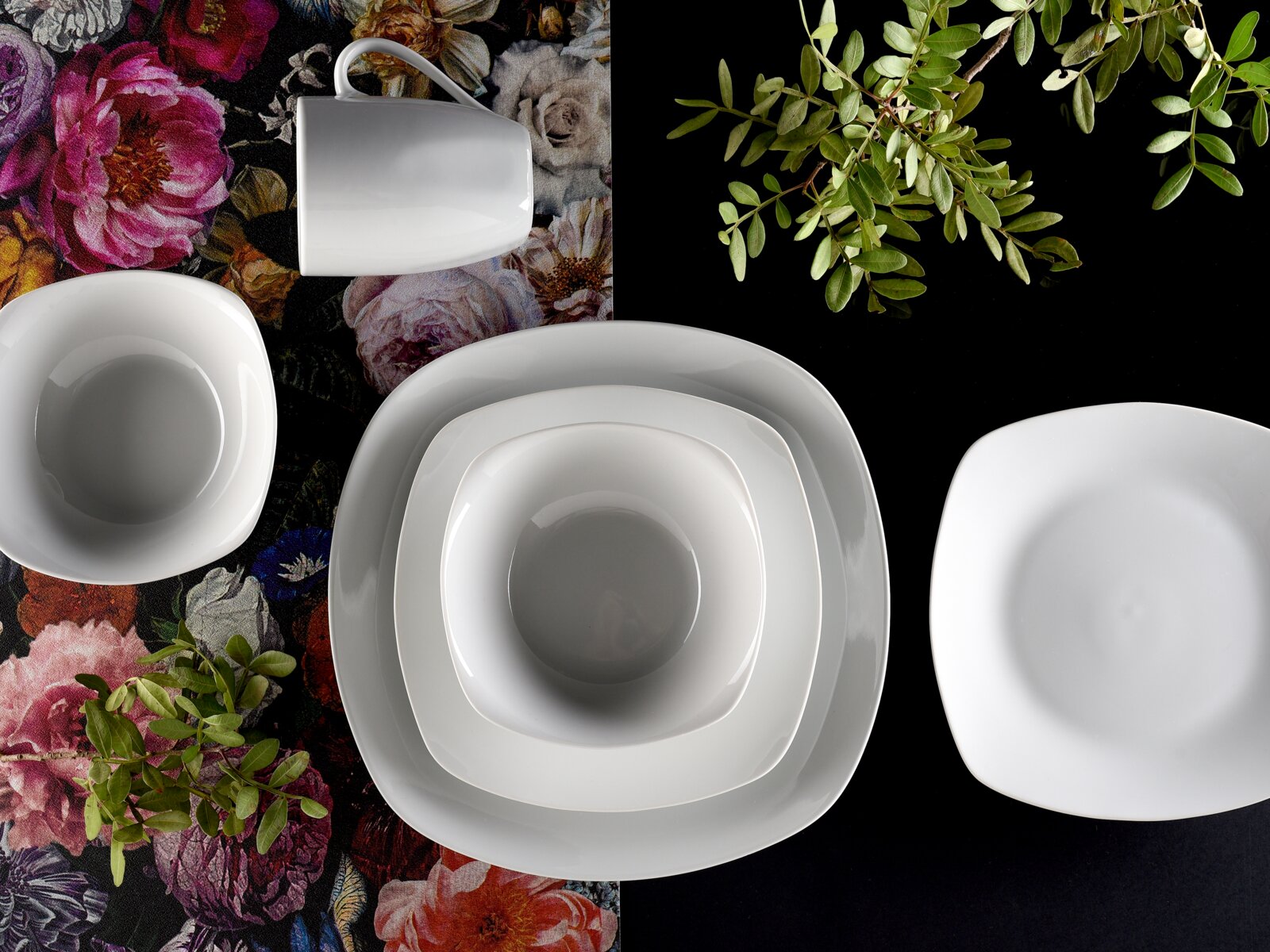 Weißes, quadratisches Porzellan-Geschirrset mit Tasse, Schale und Tellern auf floralem und schwarzem Untergrund, Draufsicht von oben.