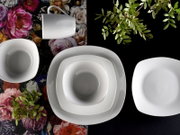 Weißes, quadratisches Porzellan-Geschirrset mit Tasse, Schale und Tellern auf floralem und schwarzem Untergrund, Draufsicht von oben.