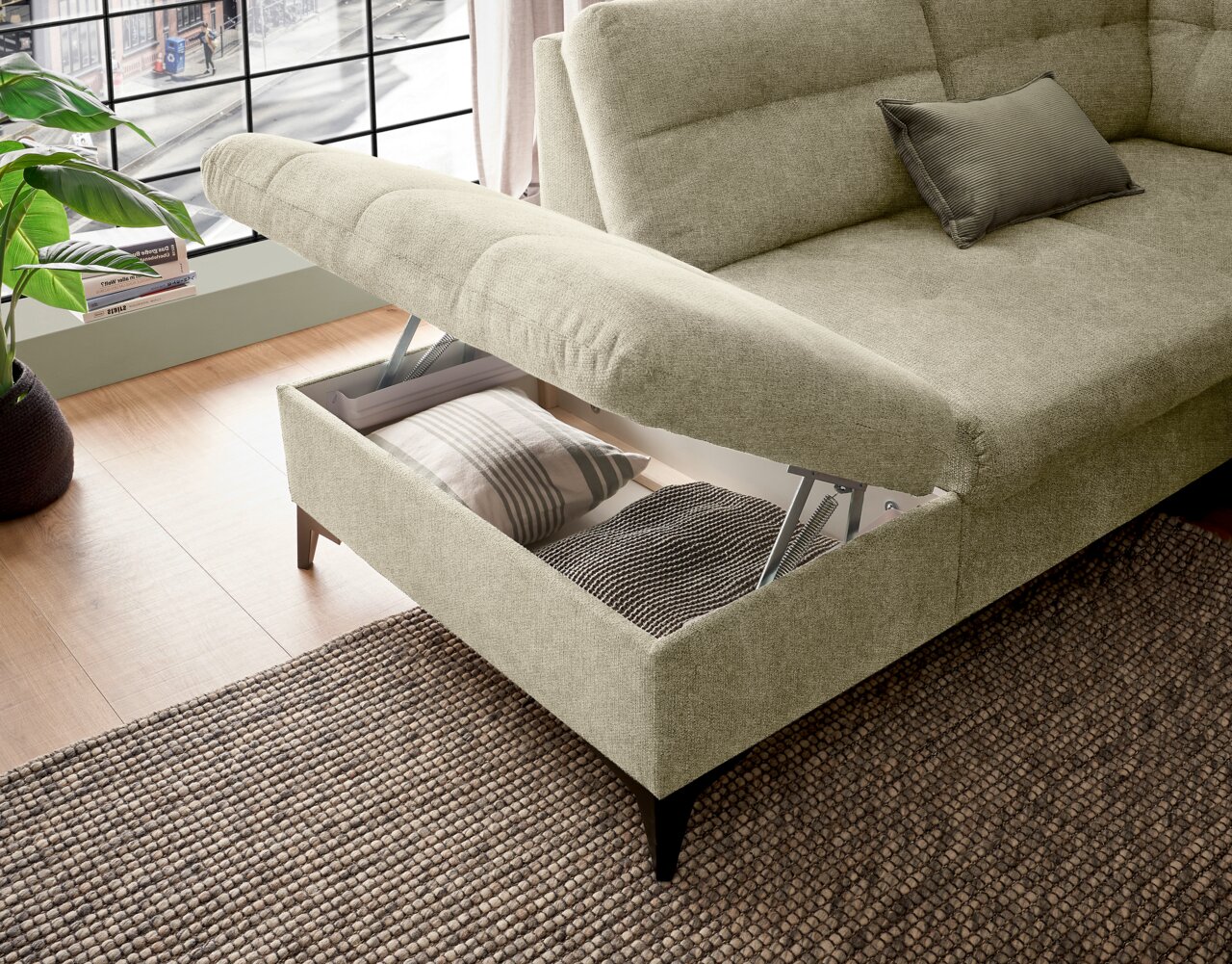 Ecksofa mit Stauraum, geöffnet, aus seitlicher Perspektive, in einem modernen Wohnzimmer mit Holzboden und großem Fenster.
