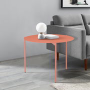 Runder Beistelltisch in Korallenfarbe, seitliche Perspektive, neben einem grauen Sofa, dekoriert mit einer Lampe und Büchern.