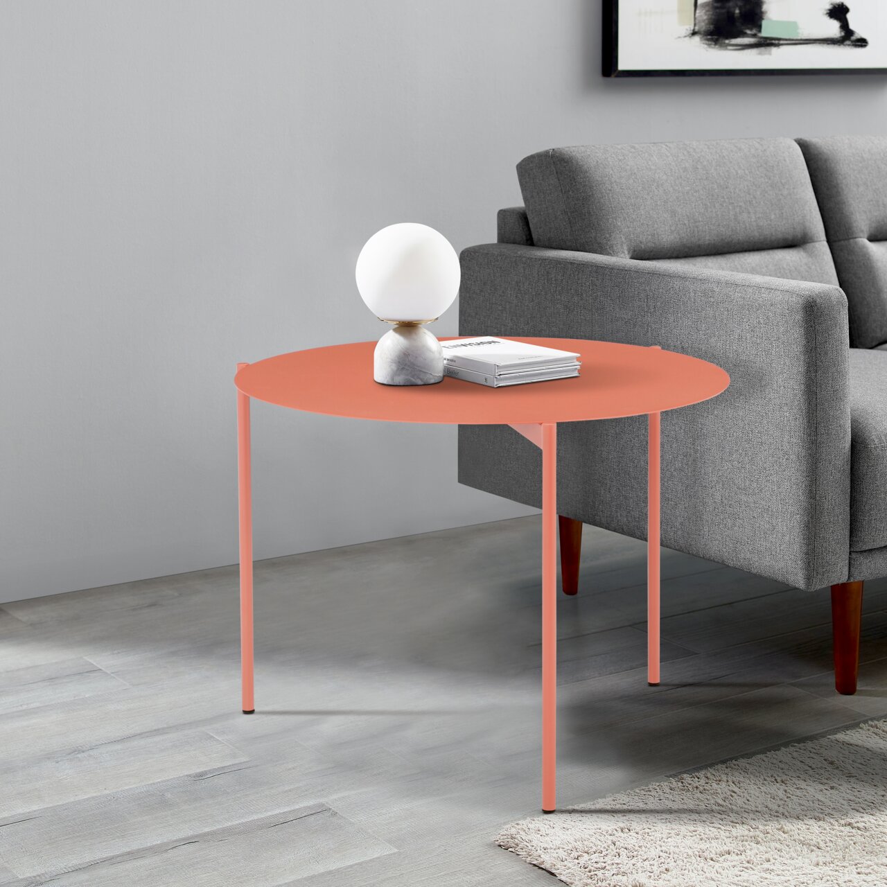 Runder Beistelltisch in Korallenfarbe, seitliche Perspektive, neben einem grauen Sofa, dekoriert mit einer Lampe und Büchern.
