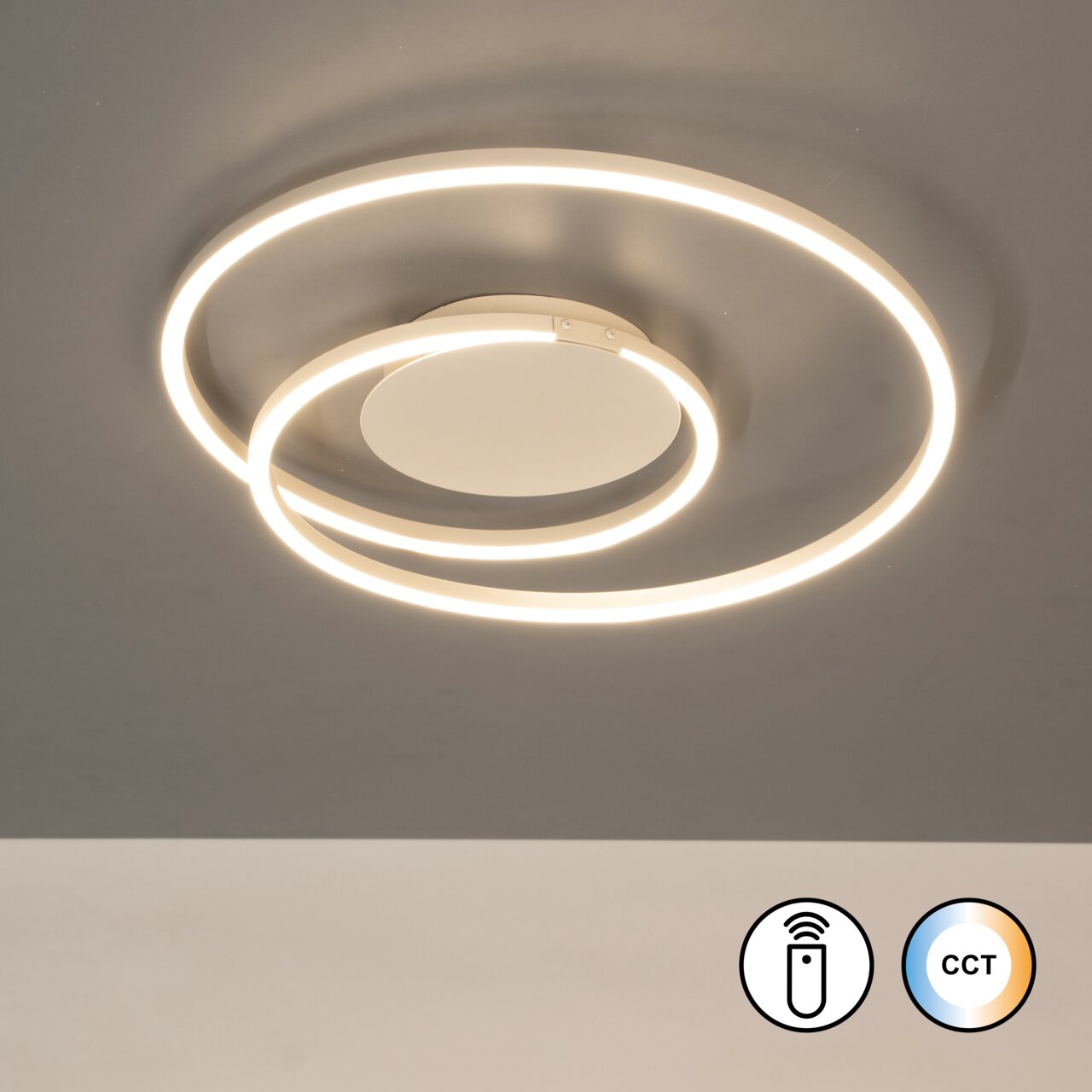 Moderne led-plafondlamp met twee verlichte ringen in warmwit, bovenaanzicht van onderaf op de aan het plafond gemonteerde lamp; symbolen voor de afstandsbediening en de CCT-kleurregeling