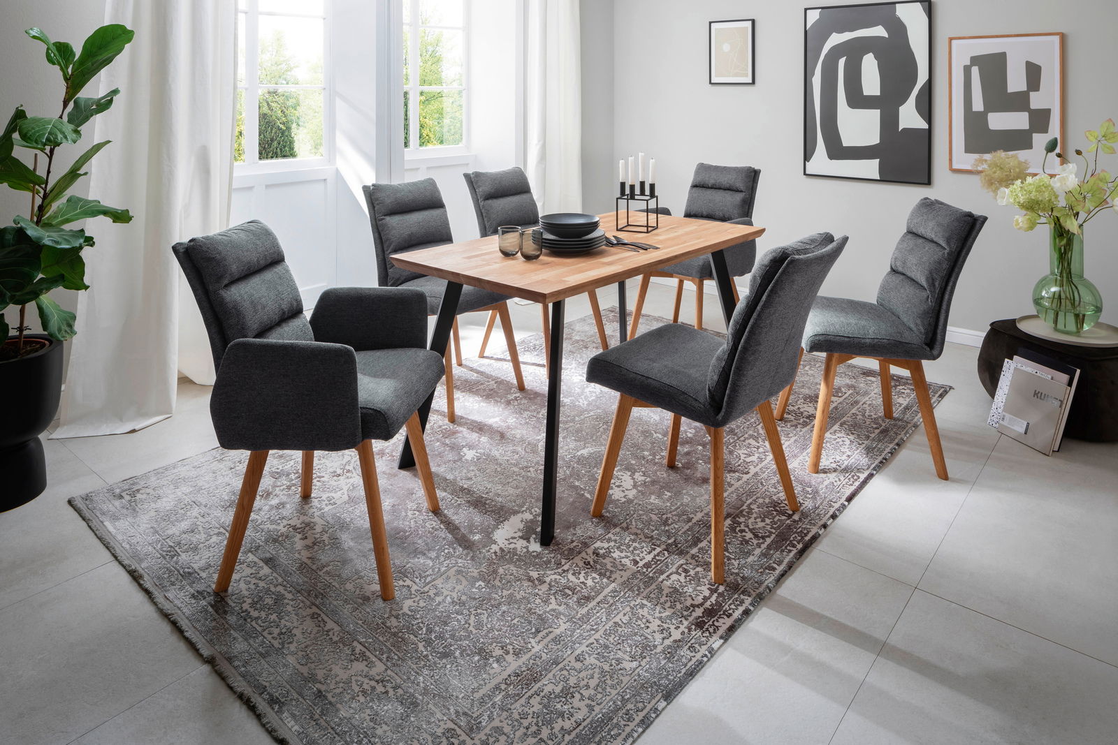Moderner Esstisch mit Holzplatte und schwarzen Metallbeinen, umgeben von sechs grauen Polsterstühlen auf einem gemusterten Teppich in einem hellen Raum. Perspektive: seitlich von vorne.
