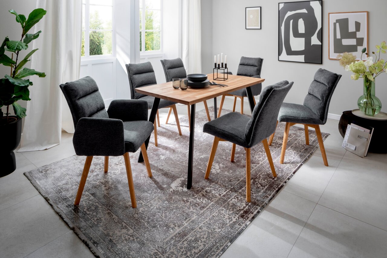 Moderner Esstisch mit Holzplatte und schwarzen Metallbeinen, umgeben von sechs grauen Polsterstühlen auf einem gemusterten Teppich in einem hellen Raum. Perspektive: seitlich von vorne.