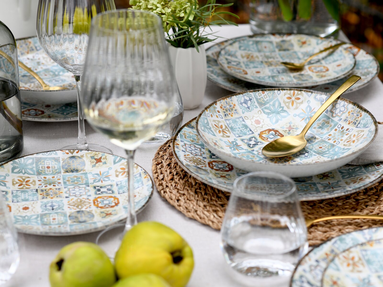 Schräg von oben fotografiertes, gedecktes Tischarrangement mit mediterran gemustertem Geschirr in Blau- und Orangetönen, goldfarbenem Besteck, Weingläsern und Wasserbechern auf hellem Tischtuch mit geflochtenem Platzset und grünen Früchten im Vordergrund.