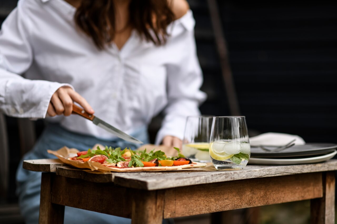 SCHOTT ZWIESEL Trinkglas PRIZMA Eine Person schneidet eine Pizza auf einem Holztisch, daneben stehen zwei Gläser mit Getränken und Eiswürfeln. Perspektive von vorne.