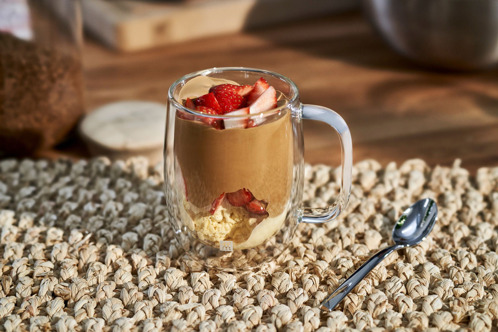 Kaffeeglas mit Griff, gefüllt mit Dessert und Erdbeeren, auf einem Tisch mit Löffel, seitliche Perspektive