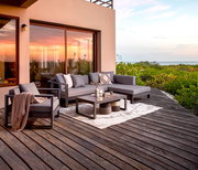 Moderne Outdoor-Lounge mit grauem Ecksofa, passendem Sessel und niedrigem Couchtisch auf Holzterrasse am Meer bei Sonnenuntergang, Perspektive: leicht erhöhte Frontansicht schräg von links