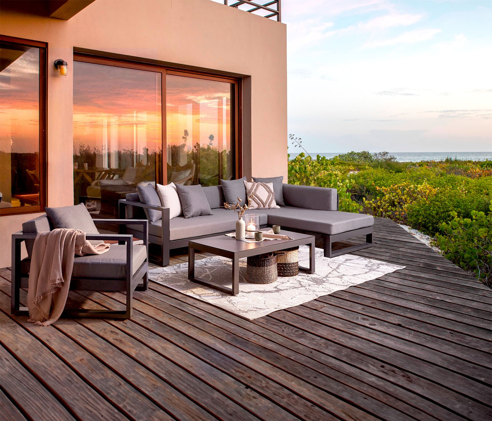 Amatio Lounge-Set (rechts) WILLIS Moderne Outdoor-Lounge mit grauem Ecksofa, passendem Sessel und niedrigem Couchtisch auf Holzterrasse am Meer bei Sonnenuntergang, Perspektive: leicht erhöhte Frontansicht schräg von links