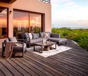 Moderne Outdoor-Lounge mit grauem Ecksofa, passendem Sessel und niedrigem Couchtisch auf Holzterrasse am Meer bei Sonnenuntergang, Perspektive: leicht erhöhte Frontansicht schräg von links