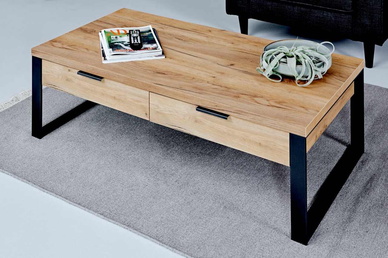 Moderner Couchtisch aus Holz mit schwarzen Metallbeinen, von schräg oben fotografiert, auf einem grauen Teppich stehend, mit Zeitschriften und einer Pflanze dekoriert.