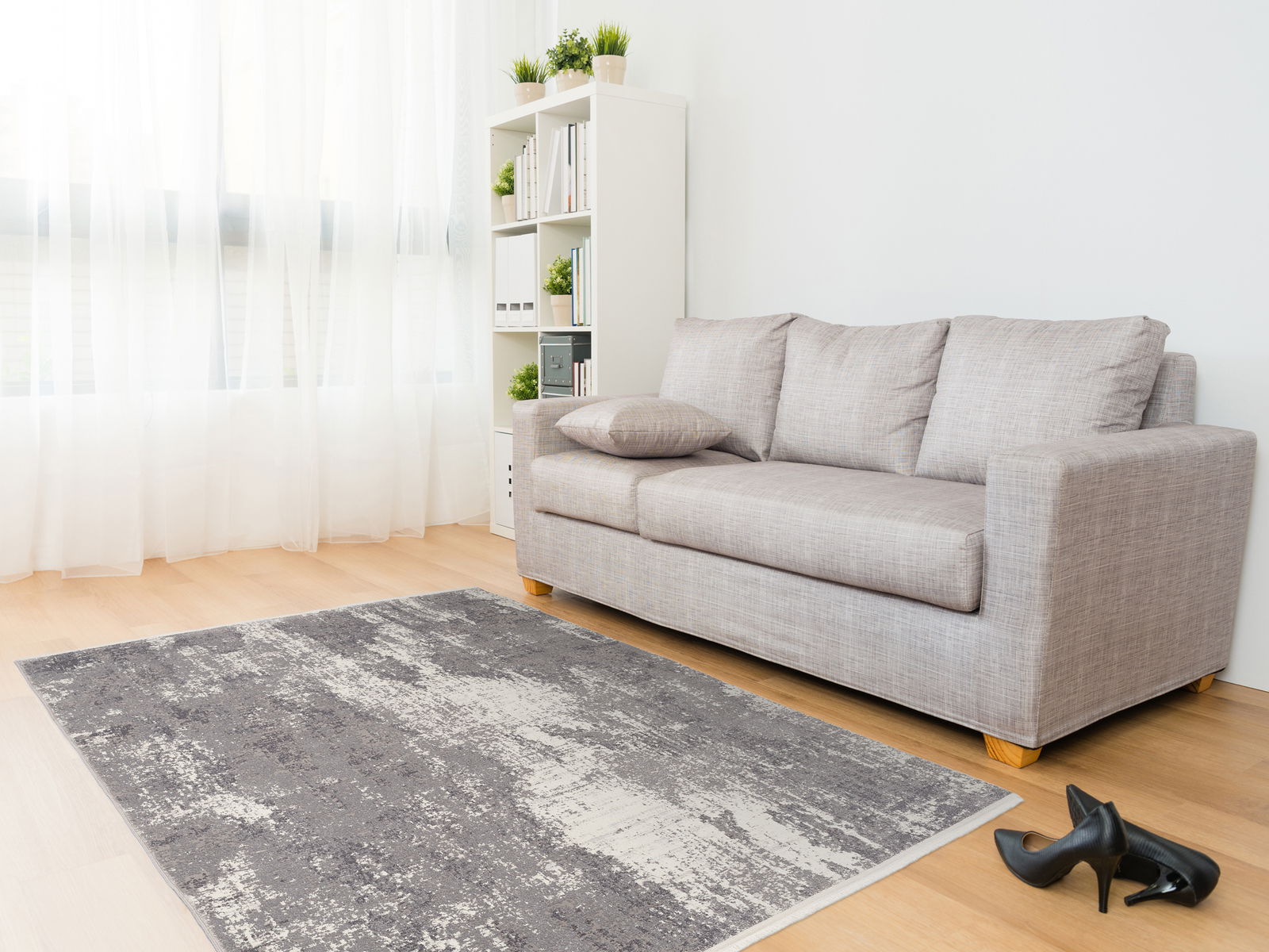 Moderner Designer Teppich in Grau- und Weißtönen auf hellem Holzboden, vor einem grauen Sofa in einem hellen Wohnzimmer mit großen Fenstern. Perspektive von schräg oben.