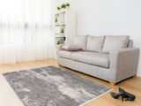 Moderner Designer Teppich in Grau- und Weißtönen auf hellem Holzboden, vor einem grauen Sofa in einem hellen Wohnzimmer mit großen Fenstern. Perspektive von schräg oben.
