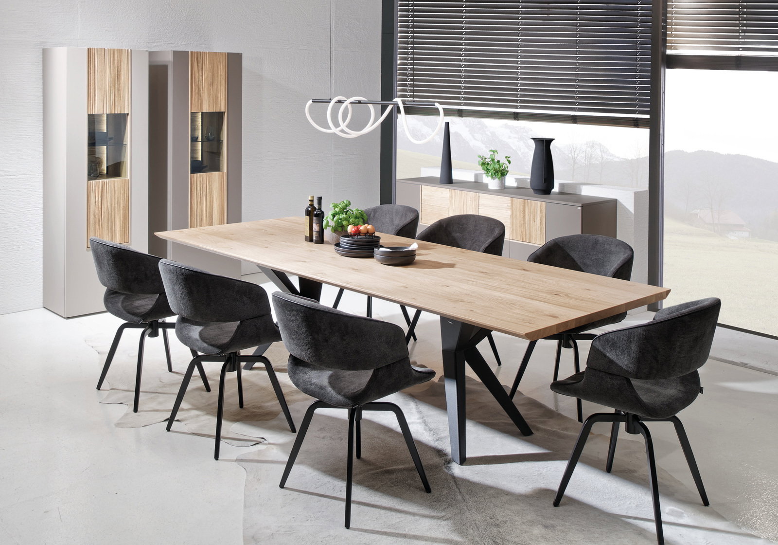 Moderner Esstisch aus Holz mit schwarzem Metallgestell, umgeben von acht grauen Stühlen, in einem hellen Raum mit großen Fenstern, seitliche Perspektive.