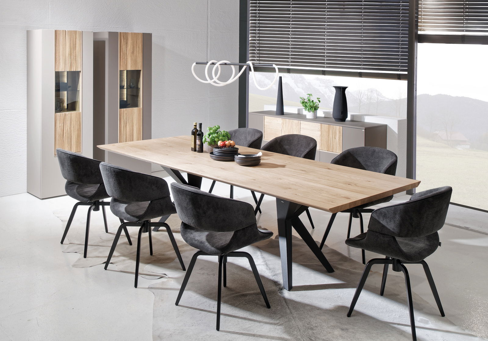 Moderner Esstisch aus Holz mit schwarzem Metallgestell, umgeben von acht grauen Stühlen, in einem hellen Raum mit großen Fenstern, seitliche Perspektive.