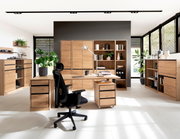 Moderne Büromöbelkombination in heller Umgebung mit Schreibtisch, Rollcontainer, Regalen und Aktenschränken aus Holz, seitliche Perspektive.
