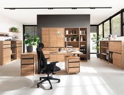 Moderne Büromöbelkombination in heller Umgebung mit Schreibtisch, Rollcontainer, Regalen und Aktenschränken aus Holz, seitliche Perspektive.